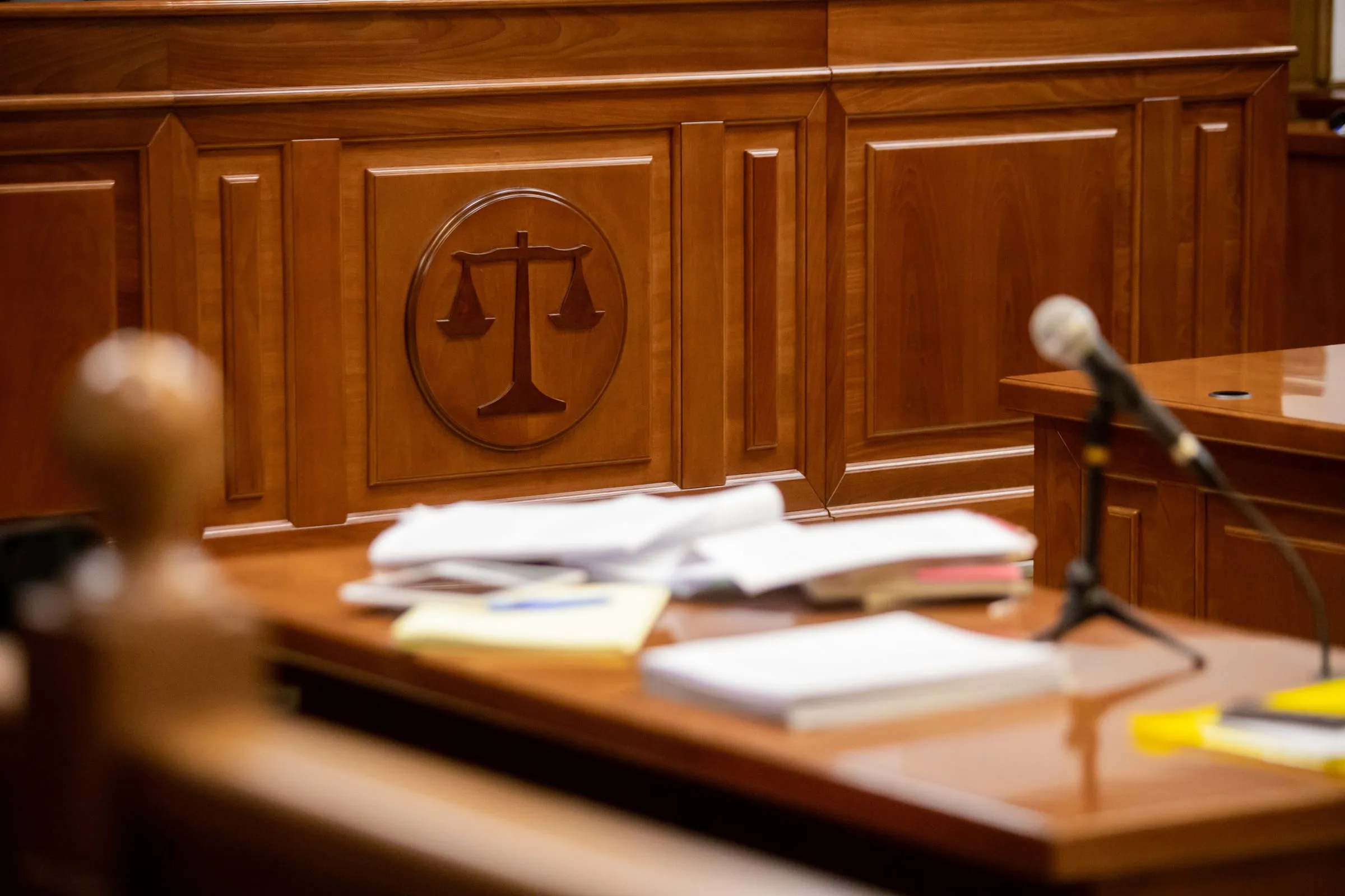 Courtroom bench with a carved scales-of-justice emblem on its wood paneling, with papers and a microphone on the desk in front